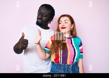 Interracial couple wearing casual clothes looking proud, smiling doing thumbs up gesture to the side Stock Photo