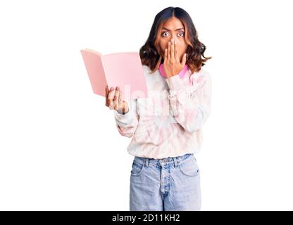 Young beautiful mixed race woman reading a book wearing headphones covering mouth with hand, shocked and afraid for mistake. surprised expression Stock Photo