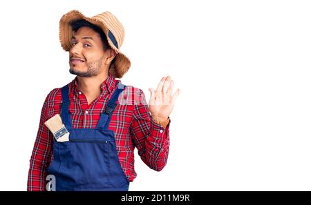 Handsome latin american young man weaing handyman uniform waiving saying hello happy and smiling, friendly welcome gesture Stock Photo