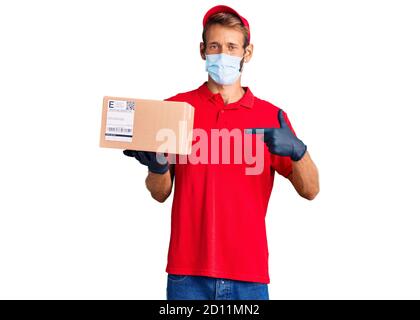 Handsome blond man with beard holding delivery box wearing medical mask smiling happy pointing with hand and finger Stock Photo