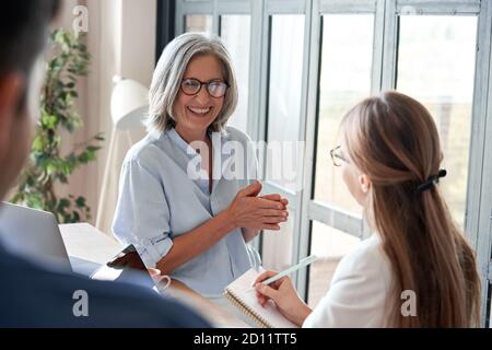 Happy old female mentor teaching young interns at group office meeting. Stock Photo