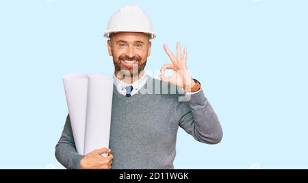 Handsome middle age man holding paper blueprints doing ok sign with fingers, smiling friendly gesturing excellent symbol Stock Photo