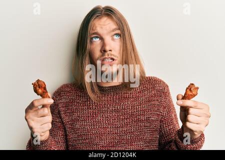 Handsome caucasian man with long hair eating chicken wings clueless and confused expression. doubt concept. Stock Photo