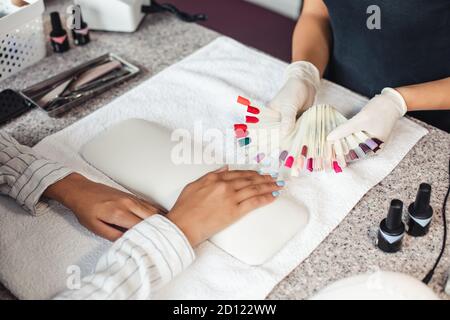 Nail polish color palette. Master in rubber gloves offers samples to african american woman client on table with manicure equipment Stock Photo