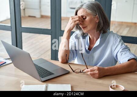 Overworked tired old lady holding glasses feeling headache after computer work. Stock Photo