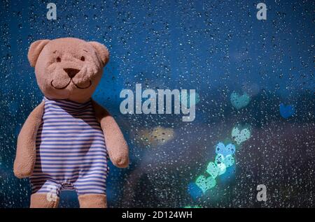 Teddy bear standing at window when raining with colorful love shape bokeh lights. Stock Photo