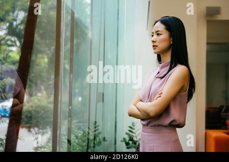 Asian businesswoman with arms folded looking through large window at work, contemplation, aspiration, ideas Stock Photo