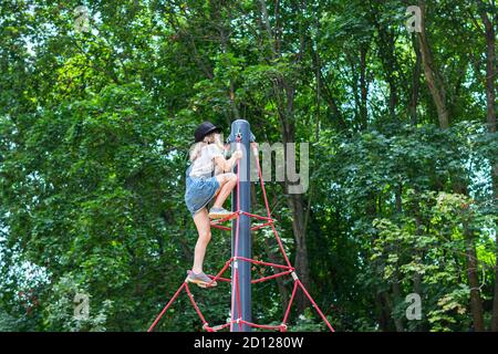 child girl in a skirt and hat climbs up the rope pyramid to the playground Stock Photo