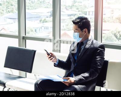 Asian businessman in suit wearing protective face mask using mobile phone while sitting in modern office building near huge glass window. Stock Photo