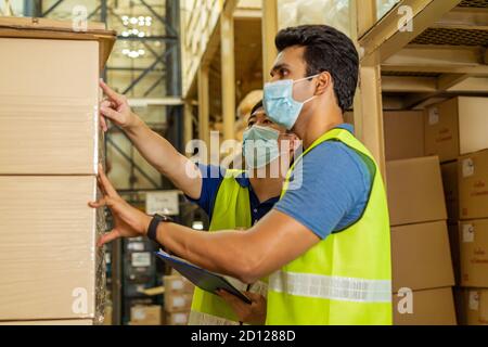 Group of young factory warehouse workers wearing a protective face mask while working in logistic industry indoor. Asian and Indian ethnic men checking item order during Coronavirus Covid 19 pandemic Stock Photo