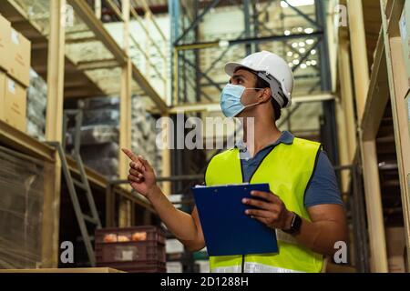 Young Indian factory warehouse worker wearing a protective face mask and safety helmet while working in logistic industry indoor. 30s man checking stock order during Coronavirus Covid 19 pandemic Stock Photo