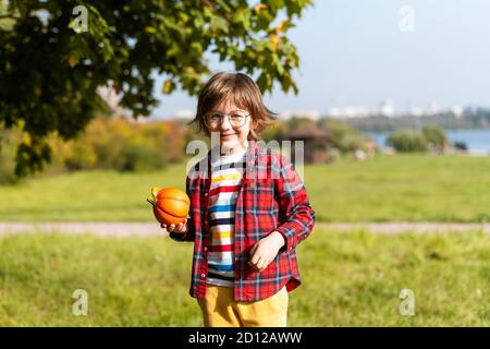 Cute boy in glasses play with pumpkin in autumn park on Halloween. Kids trick or treat. Boy carving pumpkins. Fun in fall. Dressed up child. Stock Photo