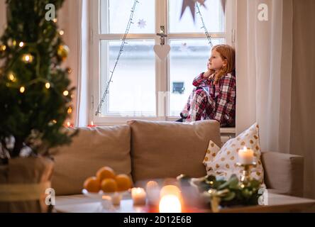 Portrait of small sad girl in pajamas indoors at home at Christmas, sitting on window.