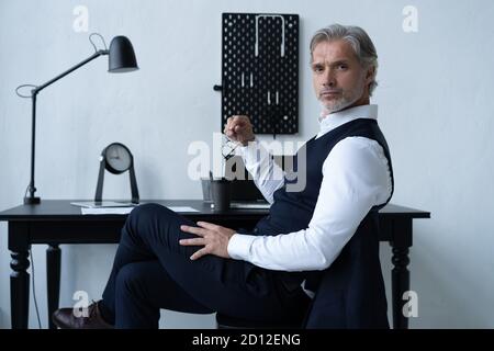 Pensive handsome matured male owner of corporation in elegant suit sitting at table while working in modern workplace. Stock Photo