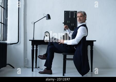 Pensive handsome matured male owner of corporation in elegant suit sitting at table while working in modern workplace. Stock Photo
