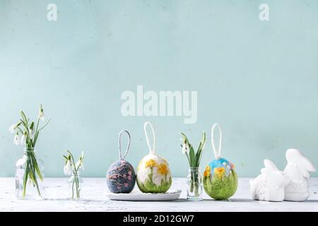 Set of craft felting Easter eggs with blooming flower sakura and green grass decor standing with spring snowdrops bouquet in glass bottle on grey tabl Stock Photo