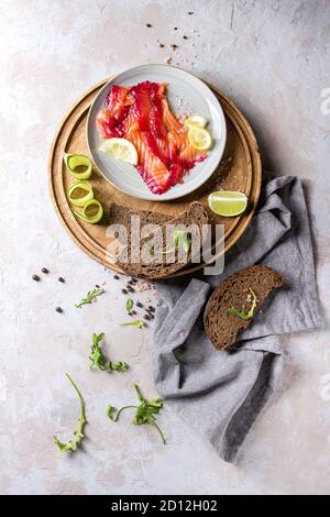 Sliced beetroot marinated salmon for sandwiches with sliced rye bread, pink salt, pepper, greens, cucumber, lemon served on wooden round tray on texti Stock Photo