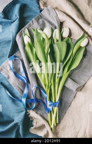 Bouquet of white spring tulips with leaves and blue ribbon over different cloth background. Top view, space. Stock Photo