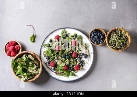 Green vegan salad with berries strawberry, blueberry, sprouts, young beetroot leaves on ceramic plate. Ingredients in wooden bowls above over grey con Stock Photo