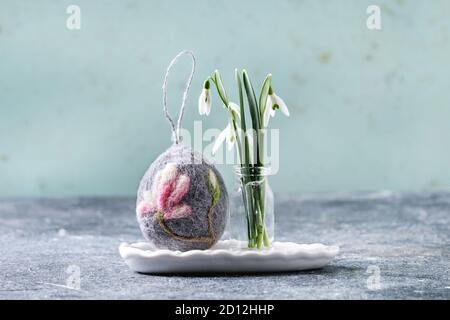 Craft felting Easter egg with blooming magnolia tree flower over grey decor standing with spring snowdrops bouquet in glass bottle on grey table. East Stock Photo
