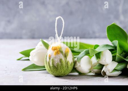 Craft felting Easter egg with blooming flower and green grass decor standing with spring tulips bouquet on grey table. Easter decoration and gifts. Co Stock Photo