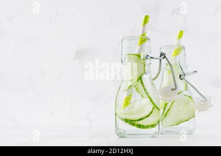 Refreshing cold detox drinks with green cucumber, bright bubbles and straw in transparent yoke bottles in elegant white interior on wood table, copy s Stock Photo