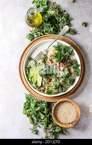 Vegan quinoa salad with kale, young beetroot leaves, garnet seeds, sliced avocado in white plate with ingredients above over grey texture background. Stock Photo