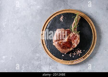 Grilled black angus beef tomahawk steak on bone served with salt, pepper and rosemary on round wooden slate cutting board over grey texture background Stock Photo