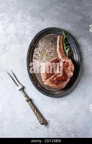 Grilled black angus beef tomahawk steak on bone served with salt, pepper and rosemary on vintage metal tray with meat fork over grey texture backgroun Stock Photo