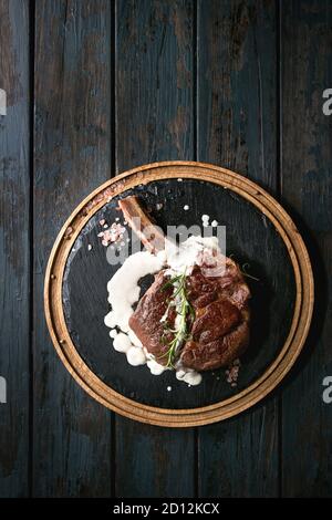 Grilled black angus beef tomahawk steak on bone served with salt, pepper, rosemary and white sauce on round wooden slate cutting board over dark woode Stock Photo