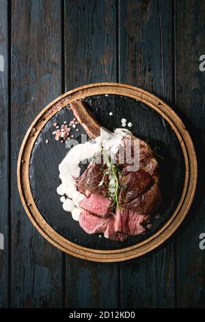 Sliced grilled sous-vide black angus beef tomahawk steak on bone served with salt, pepper, rosemary, white sauce on round slate cutting board over dar Stock Photo