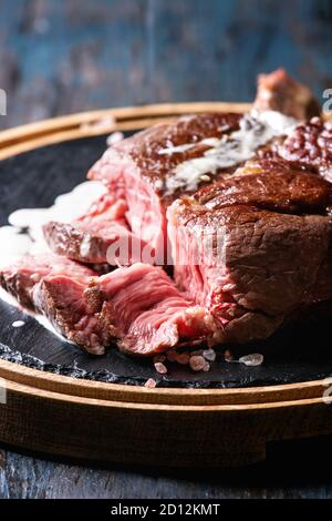 Sliced grilled sous-vide black angus beef tomahawk steak on bone served with salt, pepper, rosemary and white sauce on round wooden slate cutting boar Stock Photo