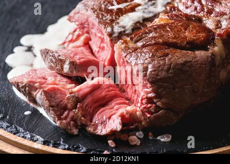 Sliced grilled sous-vide black angus beef tomahawk steak on bone served with salt, pepper, rosemary and white sauce on round wooden slate cutting boar Stock Photo