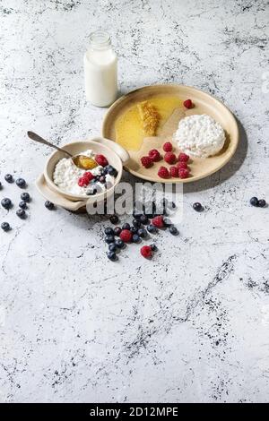 Ceramic bowl of homemade cottage cheese served with blueberries, raspberries, bottle of milk and honeycombs over white marble texture background. Copy Stock Photo
