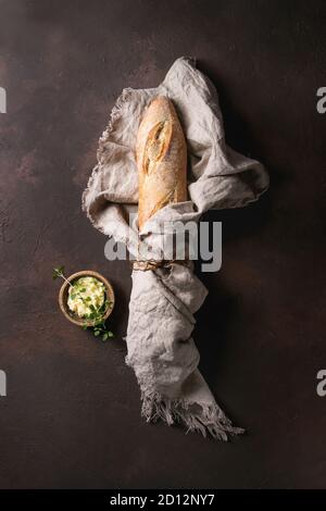 Loaf of fresh baked artisan baguette bread in linen cloth with butter and herbsover dark brown texture background. Top view, copy space. Stock Photo