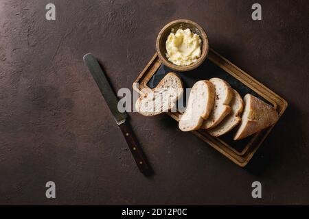 Loaf of fresh baked sliced artisan baguette bread with butter and vintage knife on wooden slate serving board over dark brown texture background. Top Stock Photo