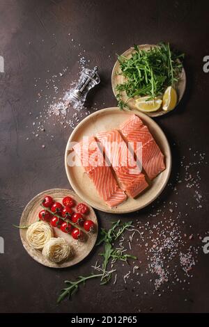 Sliced raw uncooked salmon fillet on ceramic plate with ingredients for dinner, arugula, lemon, salt, pepper, noodles, cherry tomatoes over dark brown Stock Photo