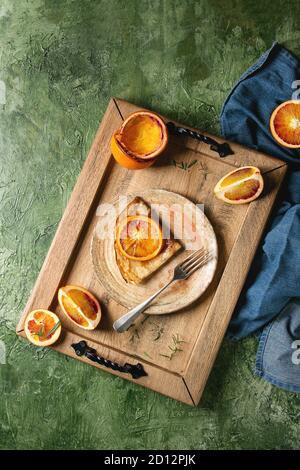 Homemade crepes pancakes served in ceramic plate with bloody oranges and rosemary syrup with sliced sicilian red oranges on wooden tray over green tex Stock Photo