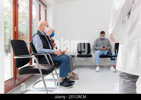 Senior couple with face masks sitting in a waiting room of a hospital together with a young man - in the foreground a physician Stock Photo