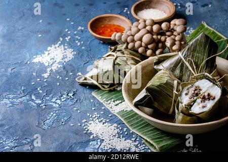 Asian rice piramidal steamed dumplings from rice tapioca flour with meat filling in banana leaves served in ceramic bowl. Ingredients and sauces above Stock Photo