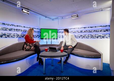 A TV show in the process of being filmed in a studio. The presenters are sitting on the studio sofa while looking at the television behind them, discu Stock Photo