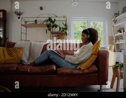 Side view of young woman relaxing at home watching webinar and typing with headphones sitting on couch Stock Photo