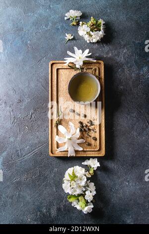 Traditional ceramic cup of hot green tea on wooden board with black iron teapot, spring flowers white magnolia and cherry blooming branches over dark Stock Photo