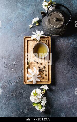 Traditional ceramic cup of hot green tea on wooden board with black iron teapot, spring flowers white magnolia and cherry blooming branches over dark Stock Photo