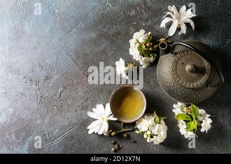Traditional ceramic cup of hot green tea with black iron teapot, spring flowers white magnolia and cherry blooming branches over dark blue texture bac Stock Photo