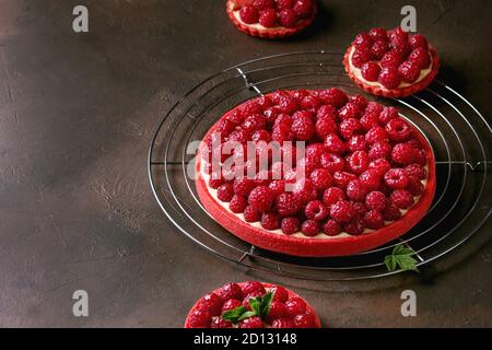 Variety of red raspberry shortbread tarts and tartlets with lemon custard and glazed fresh raspberries served on cooling rack over dark brown texture Stock Photo