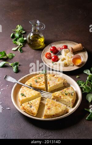 Sliced spanish potato omelette tortilla with bacon served in ceramic plate with ingredients above over dark brown texture background. Stock Photo