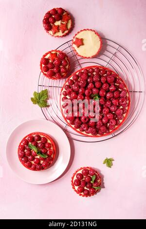 Variety of red raspberry shortbread tarts and tartlets with lemon custard and glazed fresh raspberries served on cooling rack over pink pastel backgro Stock Photo