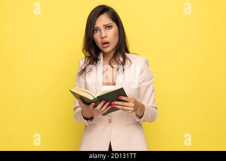 shocked young businesswoman in pink suit reading a book and making a surprised face on yellow background Stock Photo