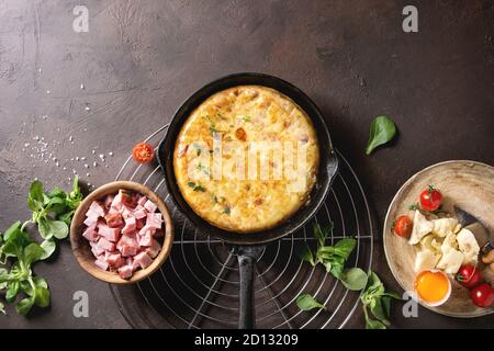 Spanish potato omelette tortilla with bacon served in cast-iron pan on cooling rack with ingredients above over over dark brown texture background. To Stock Photo